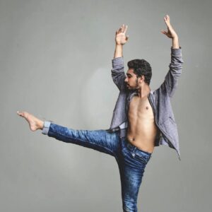 Male dancer in mid-movement, showcasing flexibility in denim jeans against a gray background.